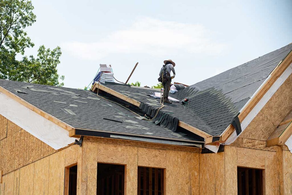 Workers remove old shingles from a house roof while protective tarps and debris containers cover the ground.