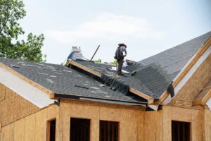 Workers remove old shingles from a house roof while protective tarps and debris containers cover the ground.